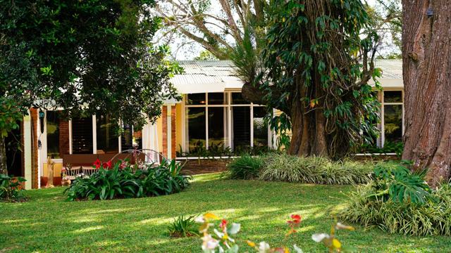 Jardín con árboles grandes y, al fondo, la casa del restaurante con ventanales.
