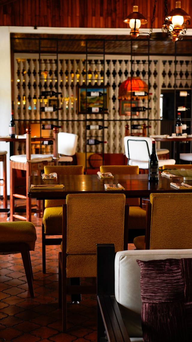 Vista vertical del salón: mesa larga con sillas amarillas, lámparas colgantes y estantes de vino al fondo.