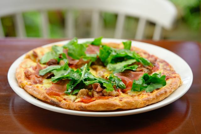 Pizza con jamón curado y hojas de arúgula en un plato blanco, sobre mesa de madera, con el jardín desenfocado al fondo.