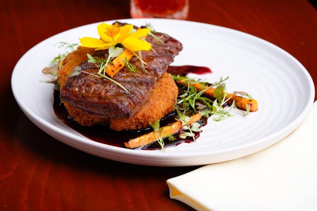 Corte de carne a la parrilla sobre una base empanizada, con zanahorias, brotes y una flor amarilla, en plato blanco.