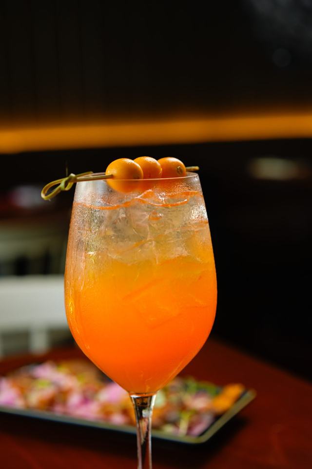 Copa con un cóctel anaranjado con hielo y tres frutas pequeñas en un palillo, sobre fondo oscuro.