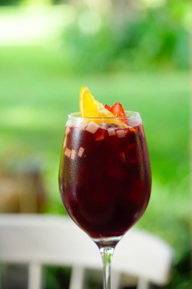 Copa de bebida tinta con frutas picadas y una rodaja de naranja, con el jardín verde desenfocado al fondo.