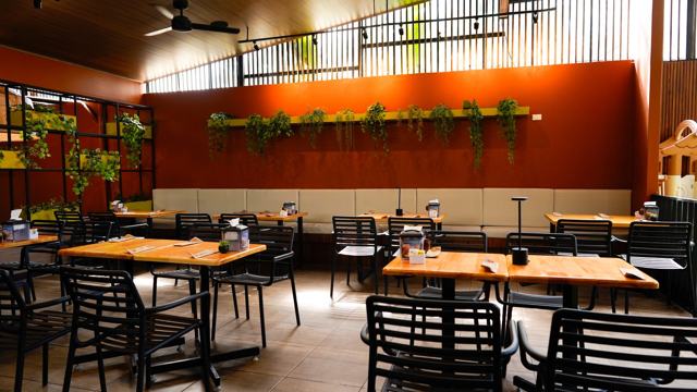 Salón con mesas de madera y sillas negras, banca corrida contra una pared terracota con plantas colgantes.