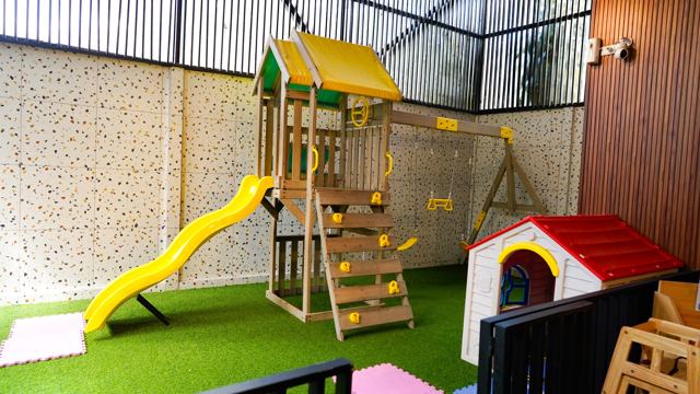 Área de juegos infantiles sobre césped sintético: torre de madera con tobogán amarillo y una casita de plástico.