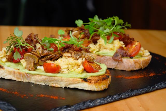 Dos tostadas con huevo revuelto, hongos, tomate y brotes, sobre una tabla de piedra negra.