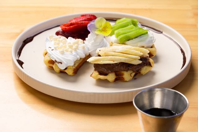 Plato con waffles cubiertos de crema batida, crema de chocolate y frutas, con un recipiente de sirope.