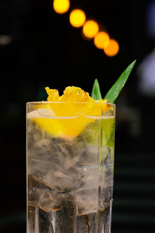 Vaso alto tallado con una bebida transparente con hielo, decorada con piña y una hoja, sobre fondo oscuro con luces.