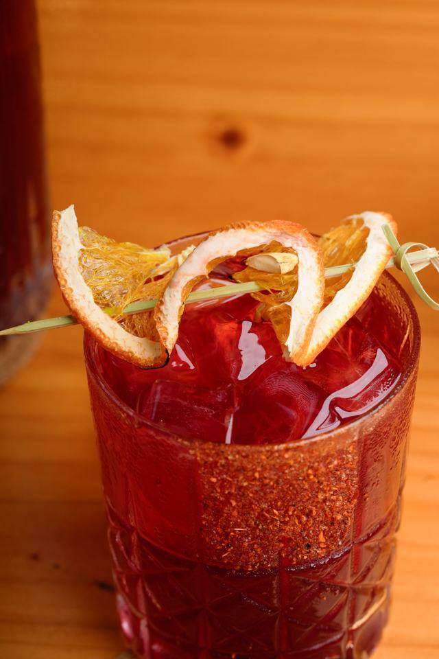 Vaso corto con una bebida roja con hielo, borde escarchado con especias y una espiral de naranja deshidratada.