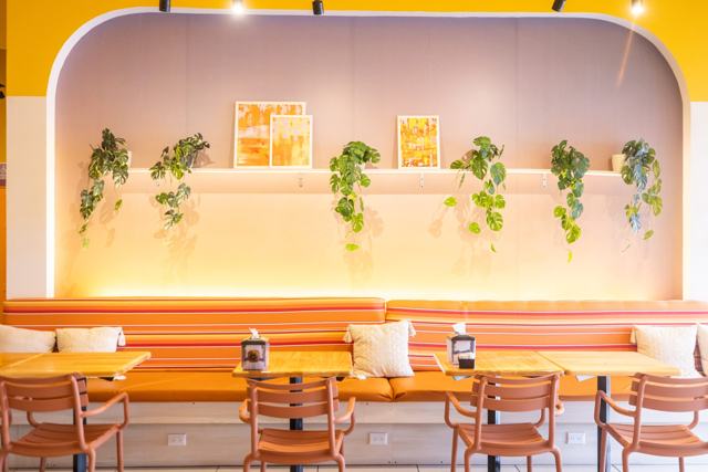 Salón con banca corrida anaranjada, mesas de madera, sillas terracota y una pared iluminada con plantas colgantes.