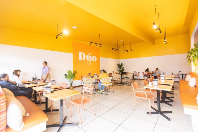 Vista amplia del salón con cielo raso amarillo, mesas y el rótulo «Dúo Café» en una pared anaranjada.