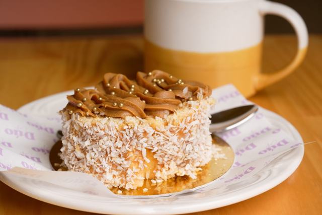 Porción de queque cubierta de coco rallado y crema de caramelo, con una taza de café al fondo.