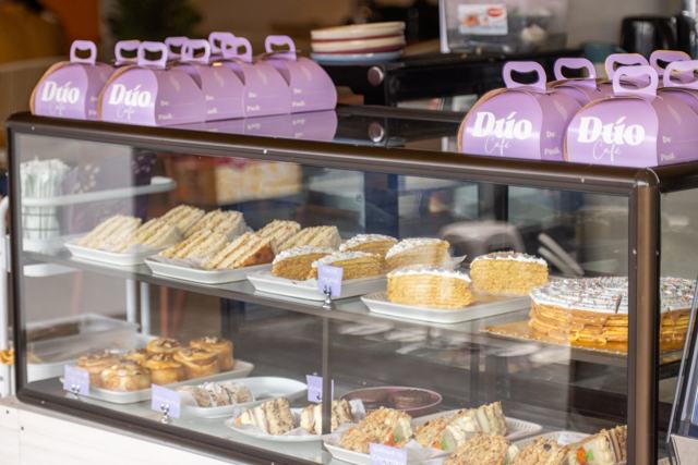 Vitrina con sándwiches, queques y repostería; encima, cajas moradas para llevar con el nombre Dúo Café.