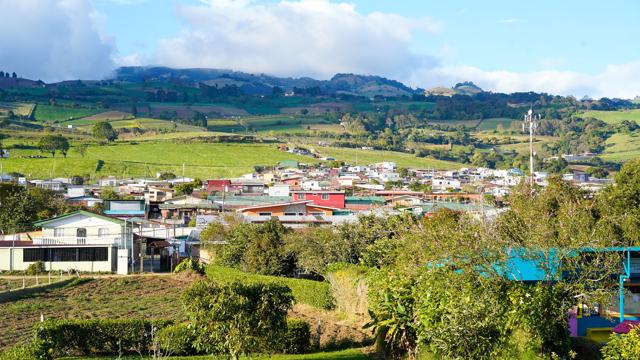 Vista de un pueblo entre lomas verdes y sembradíos, con montañas y nubes al fondo.