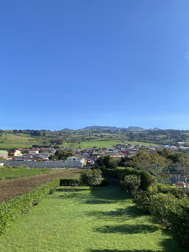 Franja de césped entre setos que baja hacia un pueblo y lomas, bajo un cielo azul despejado.