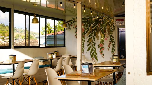Salón con mesas de madera y sillas blancas, ventanales con vista y una pared pintada con hojas y flores.
