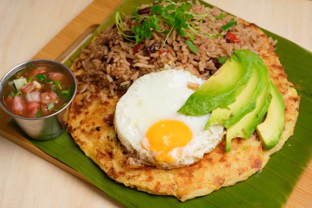 Tortilla gruesa cubierta con gallo pinto, un huevo frito y tajadas de aguacate, sobre hoja de plátano, con pico de gallo.