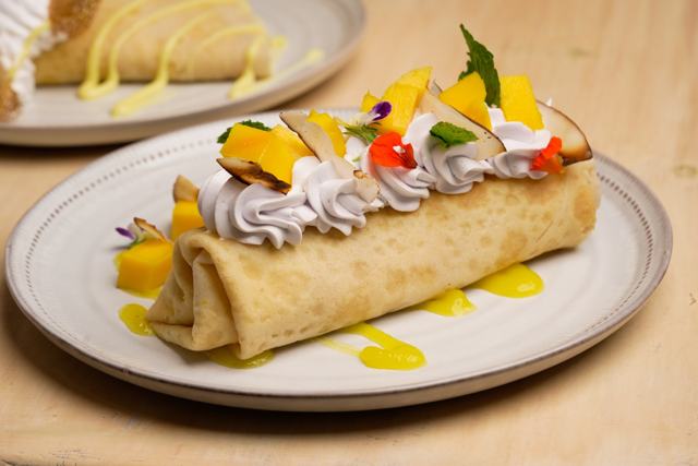 Crepa enrollada en plato blanco, cubierta con crema de taro color lila, cubos de mango, coco tostado, menta y flores comestibles, sobre salsa de mango.