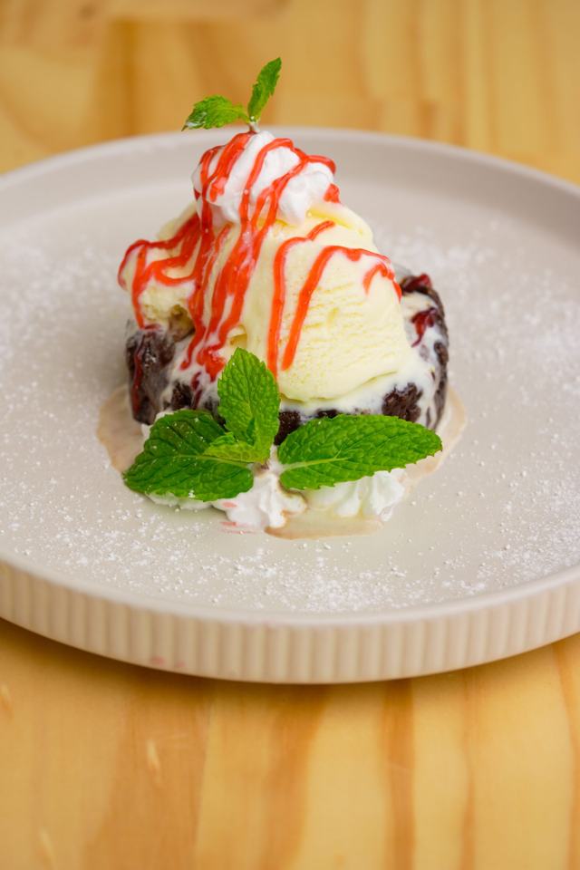 Brownie con una bola de helado, sirope rojo y hojas de menta, en plato claro.