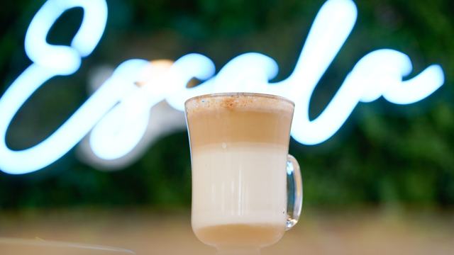 Vaso de vidrio con café con leche en capas, frente a un rótulo de neón blanco desenfocado que dice Escala.