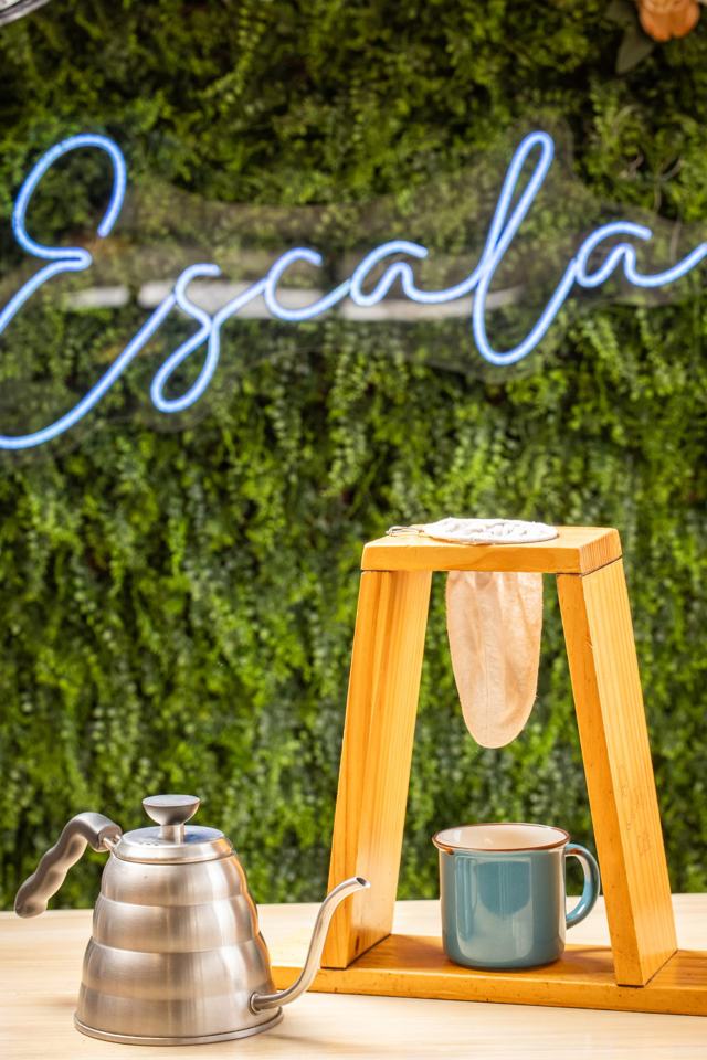Chorreador de madera con bolsa de tela, una pichel metálica y una taza, frente a un muro verde con el neón de Escala.