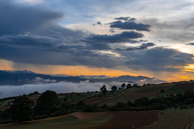 Atardecer con nubes oscuras y una franja anaranjada sobre montañas y neblina en el valle.