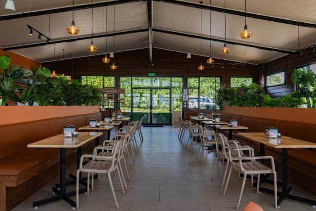 Salón amplio con techo a dos aguas, mesas de madera, sillas claras, bancas terracota y ventanales al fondo.
