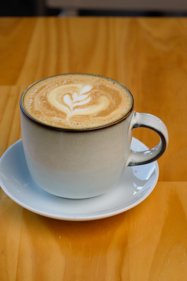 Taza de café con leche con un dibujo en la espuma, sobre plato blanco y mesa de madera.