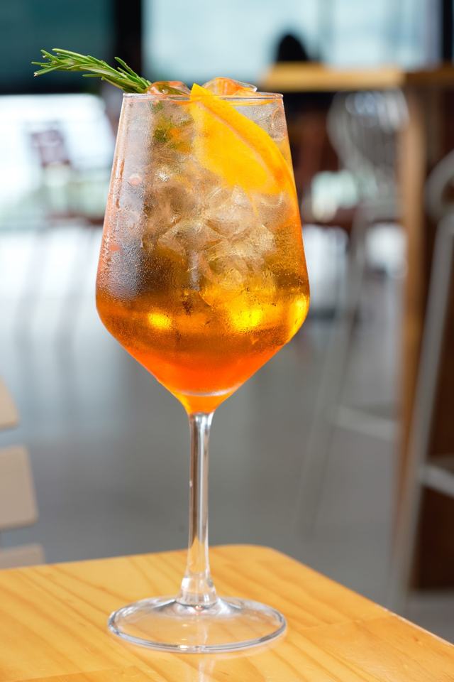 Copa con un cóctel anaranjado con hielo, rodaja de naranja y romero, con el salón desenfocado al fondo.