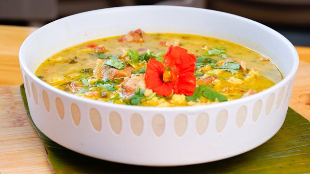 Tazón blanco de pozol con caldo amarillo, maíz, trozos de cerdo, culantro y una flor de capuchina, sobre hoja de plátano.